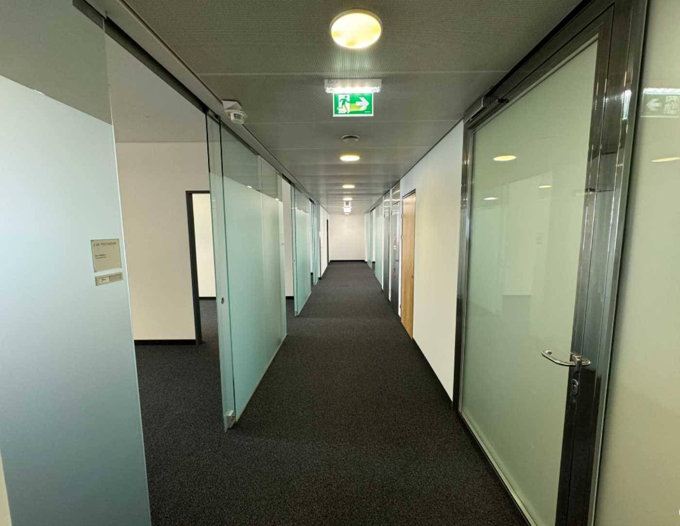 Langer Bürogang mit Teppichboden, Glaswänden und Deckenbeleuchtung, der zu weiteren Räumen führt.