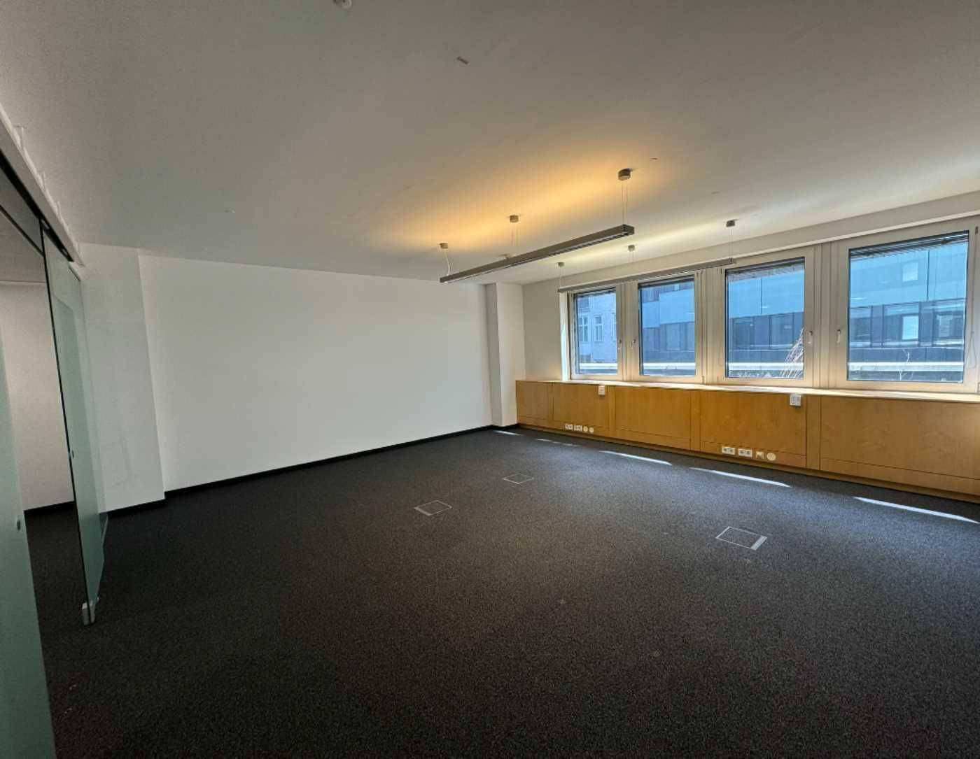 Leerer Büroraum mit Teppichboden, großen Fenstern und Holzschränken entlang der Wand.