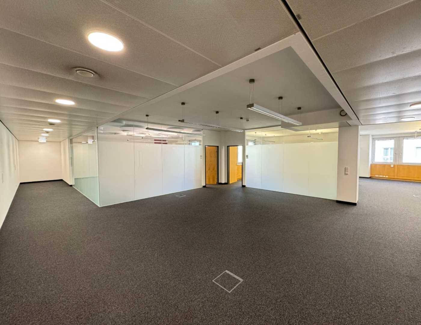 Großzügiges Büro mit Teppichboden, Glaswänden und abgehängter Decke mit integrierter Beleuchtung.
