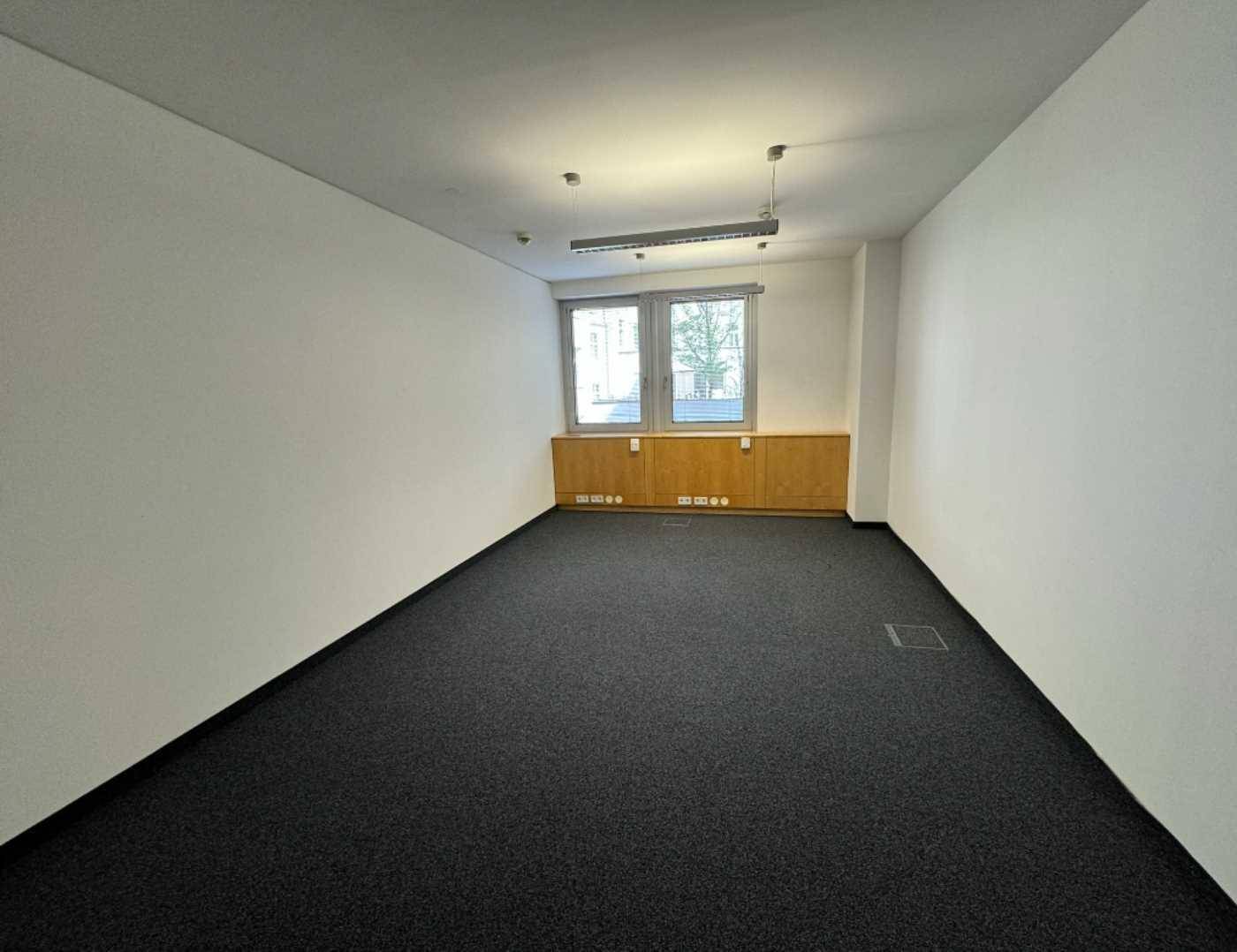 Schlichter Büroraum mit Teppichboden, weißen Wänden und einem Fenster mit Blick nach draußen.