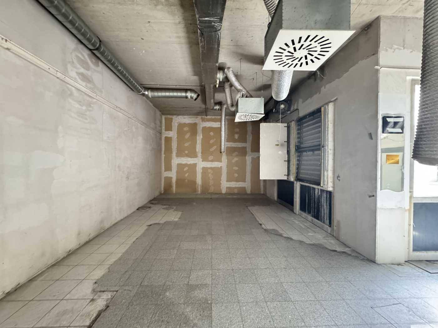 Leerer Gewerberaum mit sichtbaren Lüftungsrohren und Fliesenboden, bereit für den Ausbau.