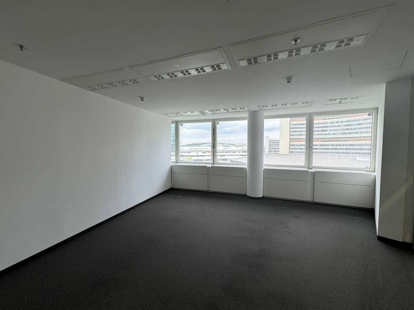 Leeres Büro mit dunklem Teppichboden und großen Fenstern, die eine helle und offene Arbeitsatmosphäre schaffen.