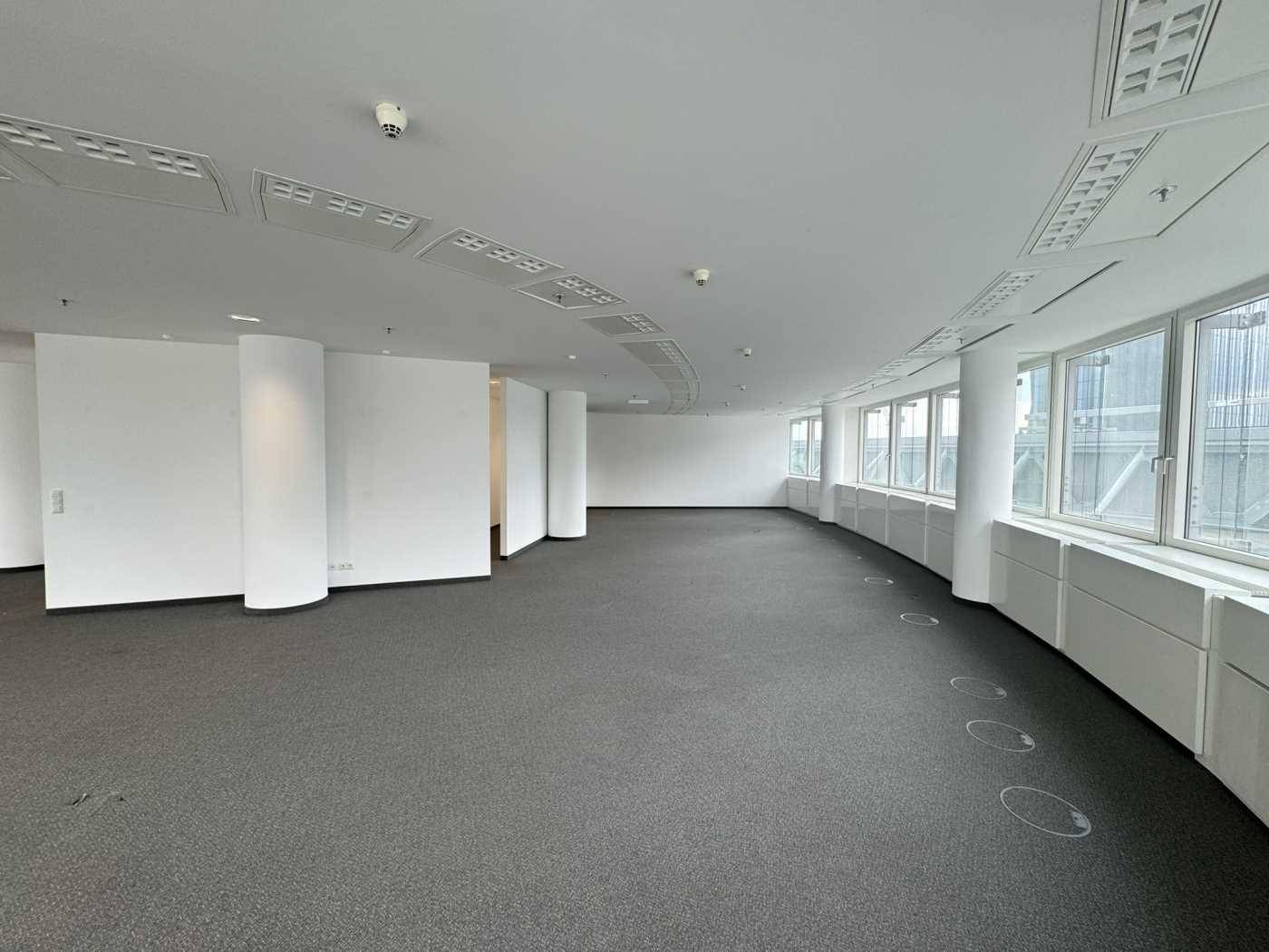 Großer, offener Bürobereich mit Teppichboden und einer geschwungenen Fensterfront, die viel Licht spendet.