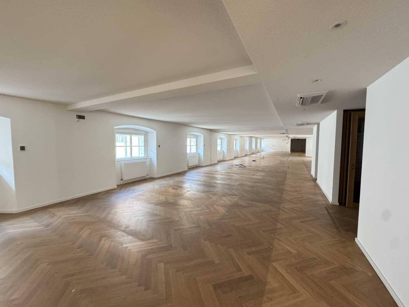 Langer, heller Büroraum mit Fischgrätparkett, vielen Fenstern und sichtbarer Klimaanlage an der Decke.