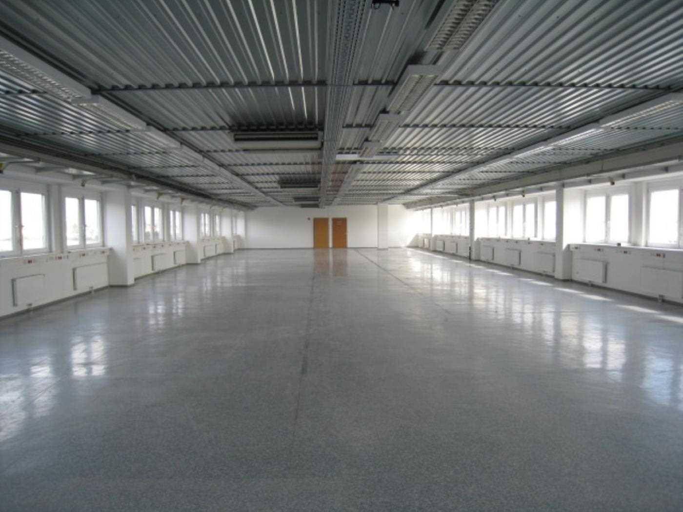 Großer, leerer Büroraum mit vielen Fenstern, grauem Boden und sichtbaren Deckeninstallationen.