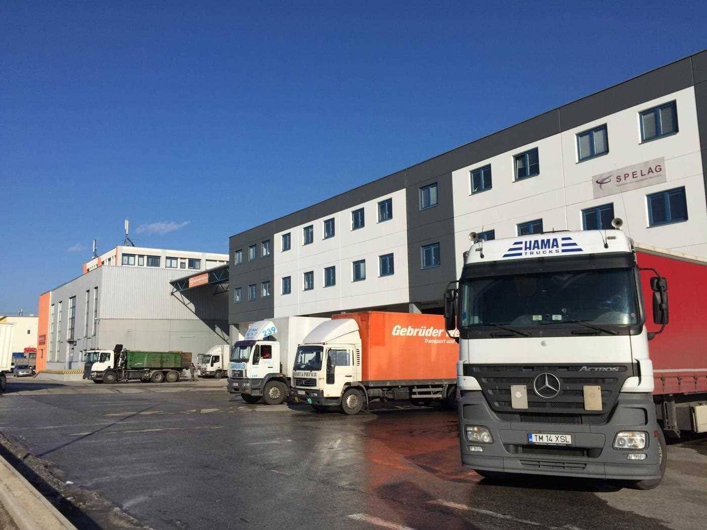 Großer Logistikbereich mit mehreren Lastwagen vor einem modernen Büro- und Lagergebäude.