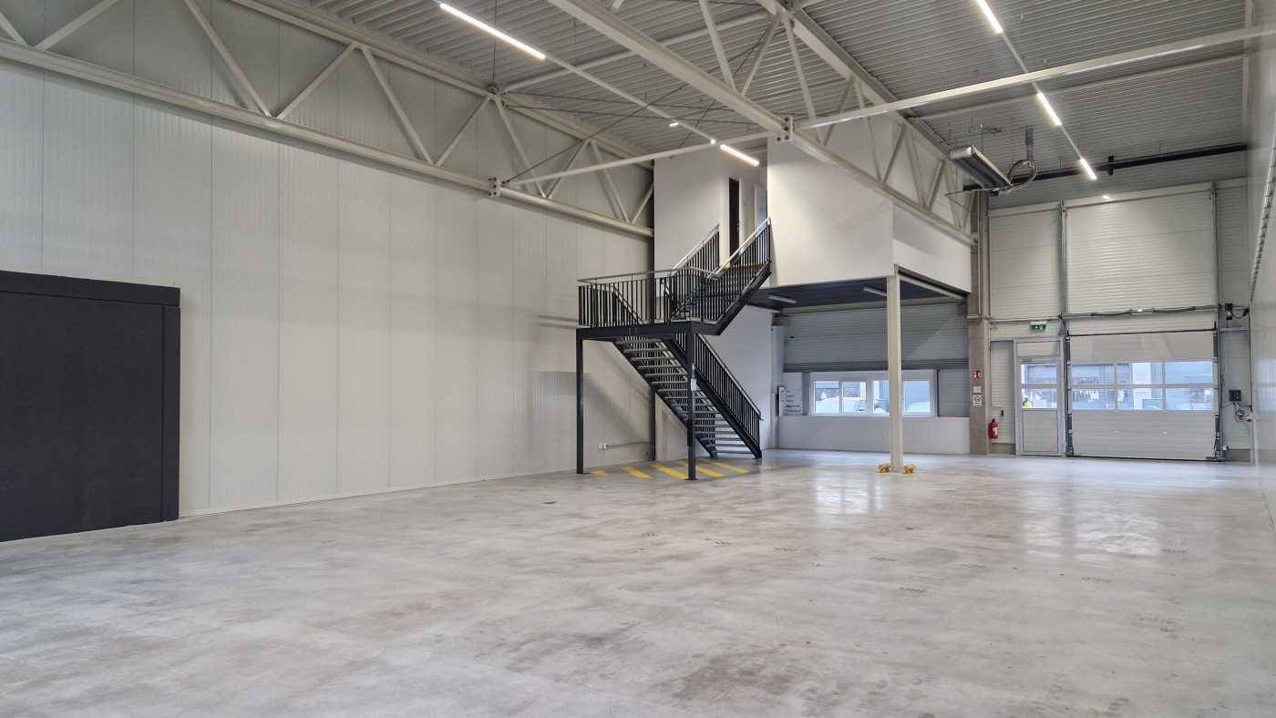 Geräumige Lagerhalle mit hohen Decken, Betonboden und einer freistehenden Stahltreppe zu einer oberen Ebene.