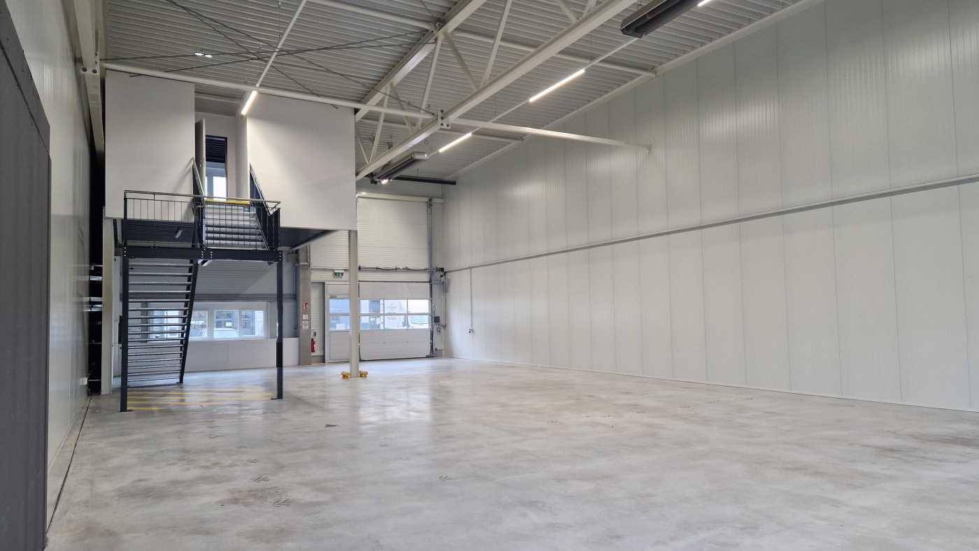 Große, helle Lagerhalle mit Betonboden, hohen Decken und einer schwarzen Stahltreppe zu einer Zwischenebene.
