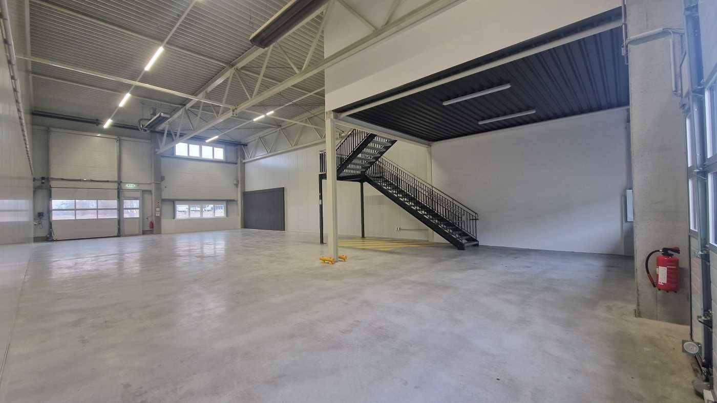 Weitläufige Lagerhalle mit Betonboden, hohen Decken und einer schwarzen Stahltreppe, die zu einer Galerie führt.