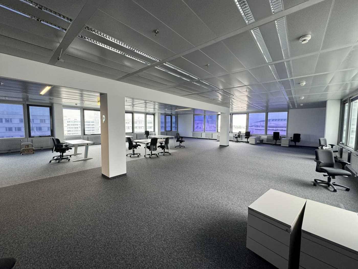 Weitläufiges Büro mit Teppichboden, vielen Fenstern und einem Blick auf die umliegenden Gebäude der Stadt.