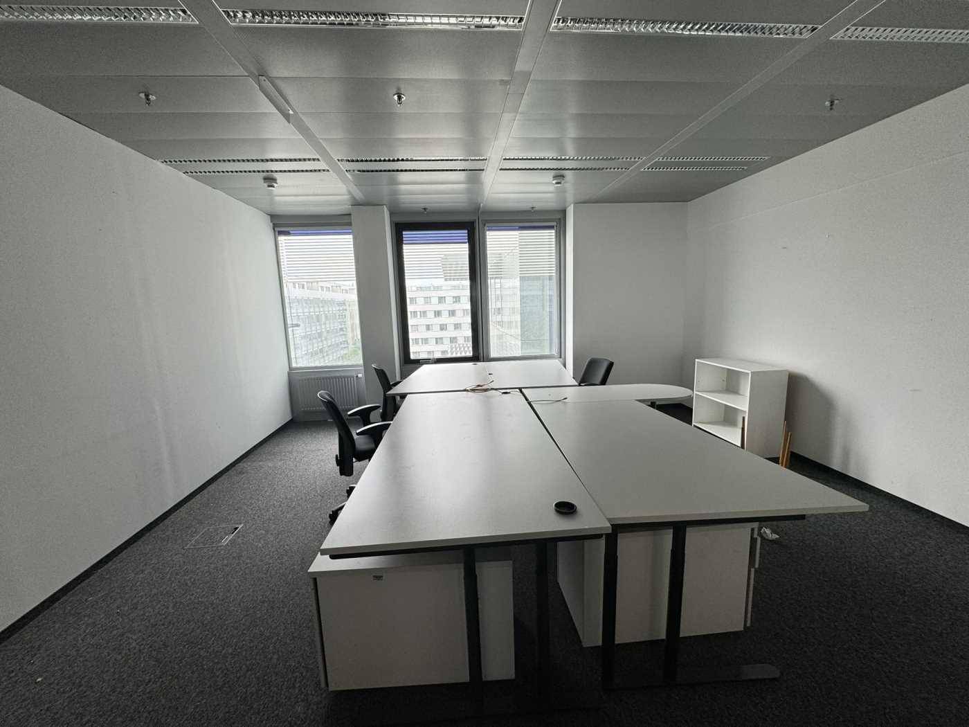 Möbliertes Büro mit Schreibtischen und Stühlen, Teppichboden und Fenstern mit Jalousien.