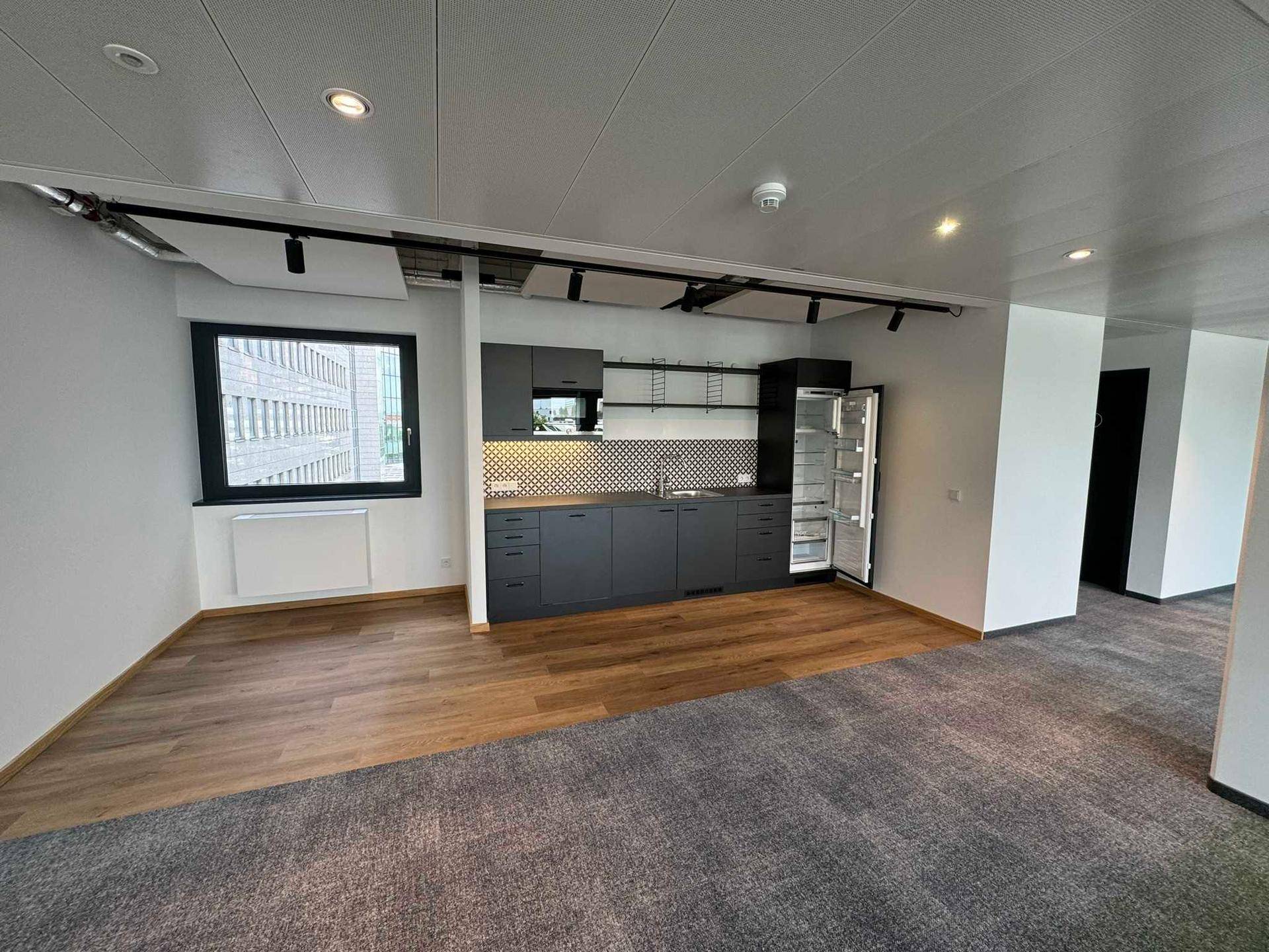 Kleine, moderne Küchenzeile in einem Büroraum mit dunklen Schränken, Kühlschrank und Holzboden.