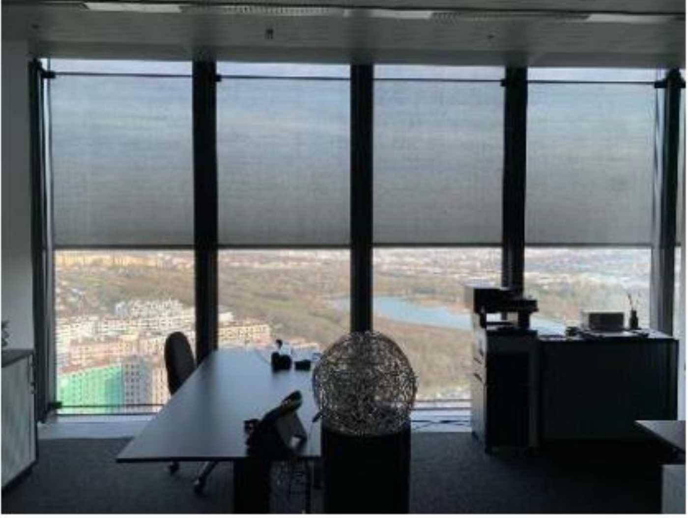 Büro mit großen Fenstern und Rollos, die einen weiten Blick auf die Stadt und einen See bieten.