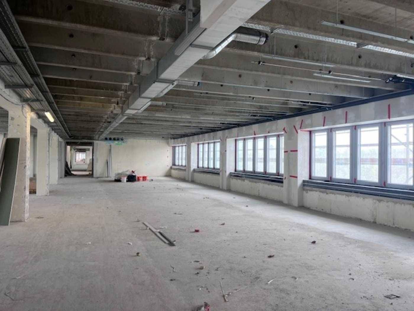 Weitläufiger, unfertiger Bürobereich mit Sichtbetondecke, großen Fenstern und offener Installation.