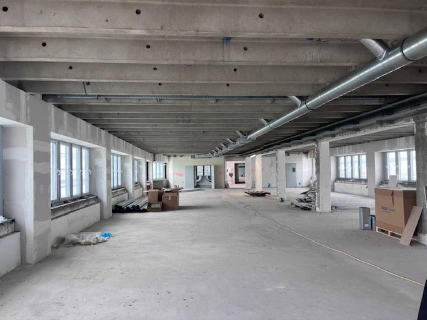 Geräumiger, unfertiger Büroraum mit Betonstruktur, vielen Fenstern und Baumaterialien.
