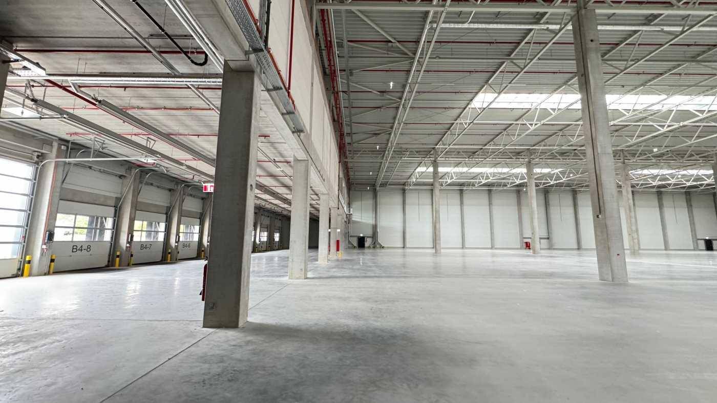 Weitläufige Lagerhalle mit hohen Decken, Betonstützen und Rolltoren im Hintergrund, sehr hell.