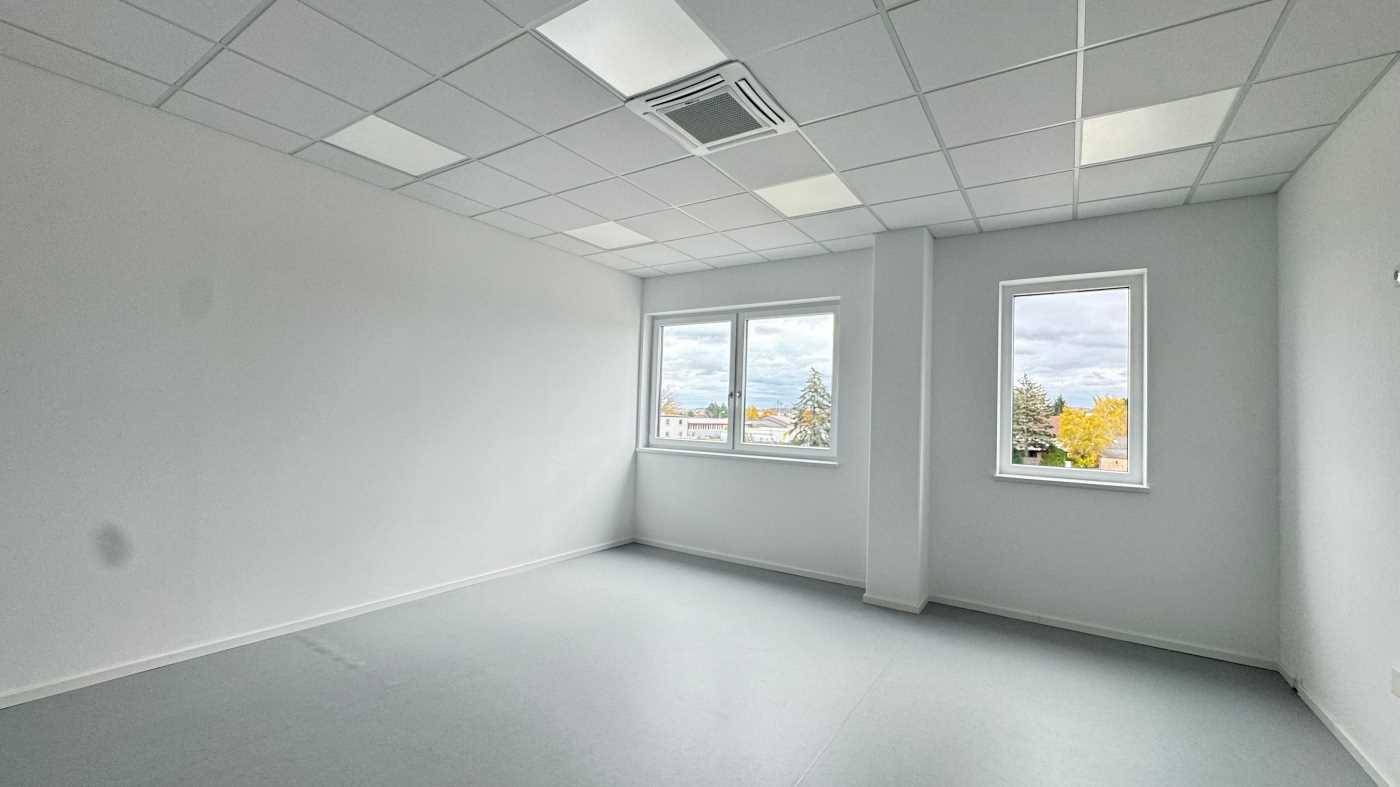 Helles Büro mit großen Fenstern, weiß gestrichenen Wänden und grauem Bodenbelag.