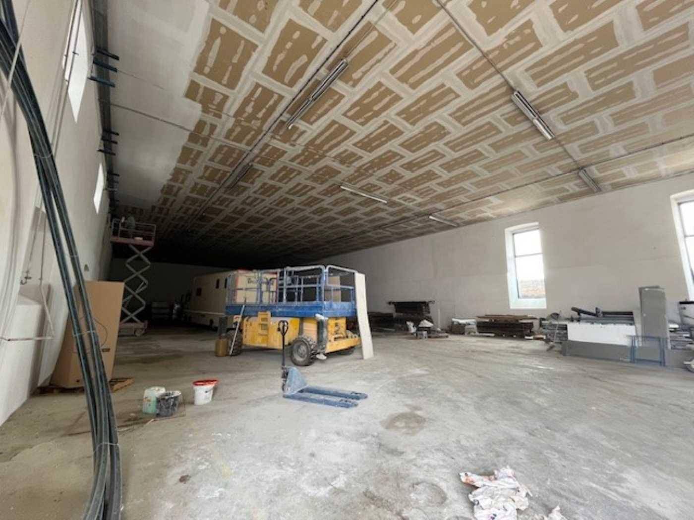 Weitläufiger Lagerbereich mit Betonboden und hohen Decken, ausgestattet mit Baumaschinen und Tageslicht.