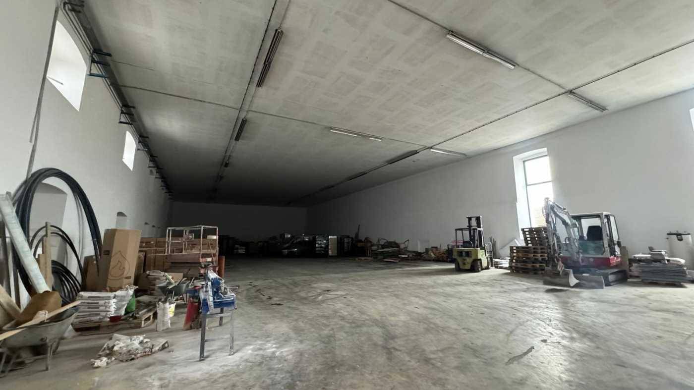 Weitläufiger Lagerraum mit Betonboden, hohen Decken und Tageslicht durch mehrere Fenster.