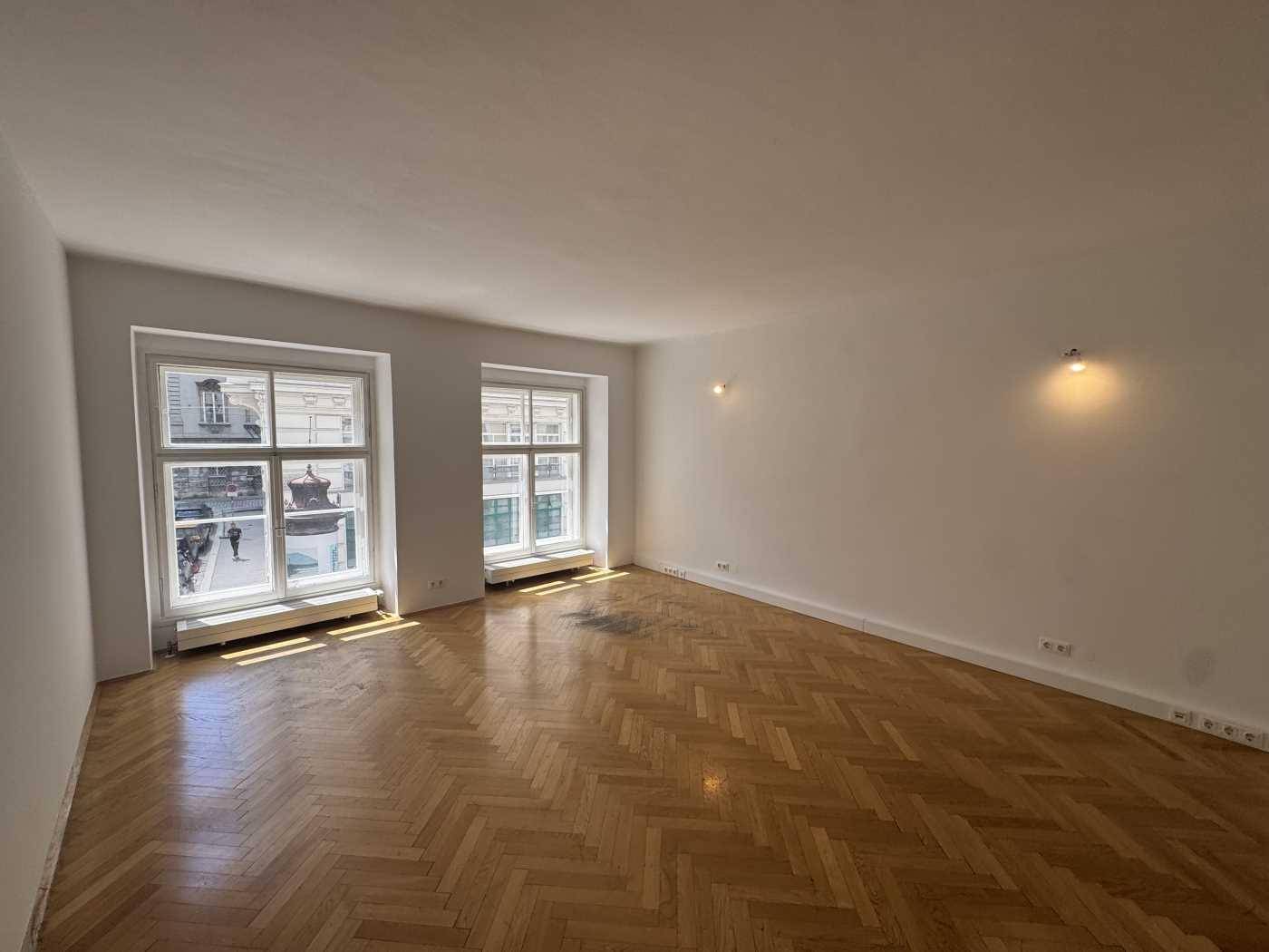 Heller Wohnraum mit klassischem Fischgrätparkett und zwei hohen Fenstern, die den Blick nach draußen freigeben.