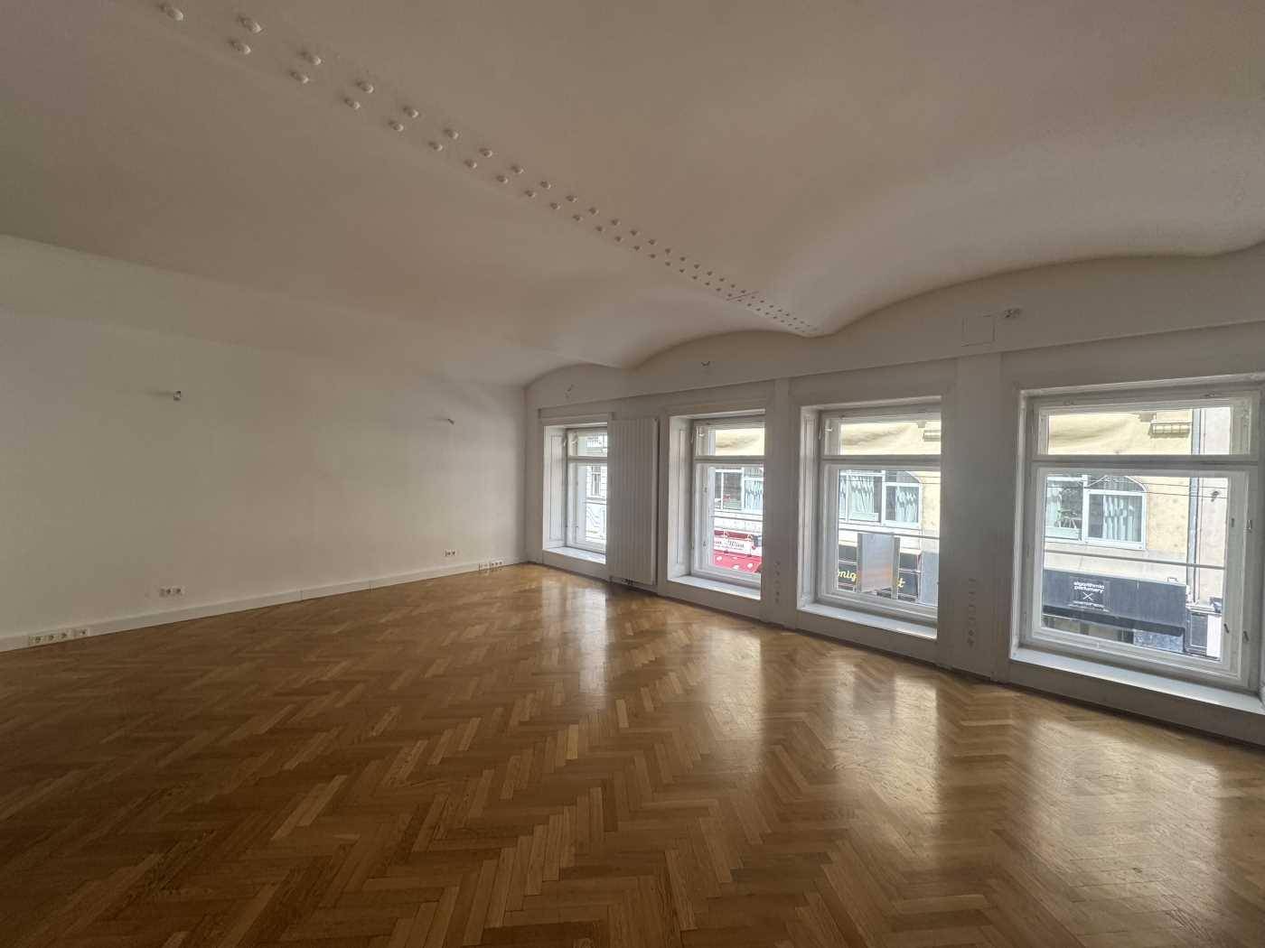 Geräumiger Raum mit glänzendem Fischgrätparkett und einer Reihe von Fenstern, die viel Licht hereinlassen.