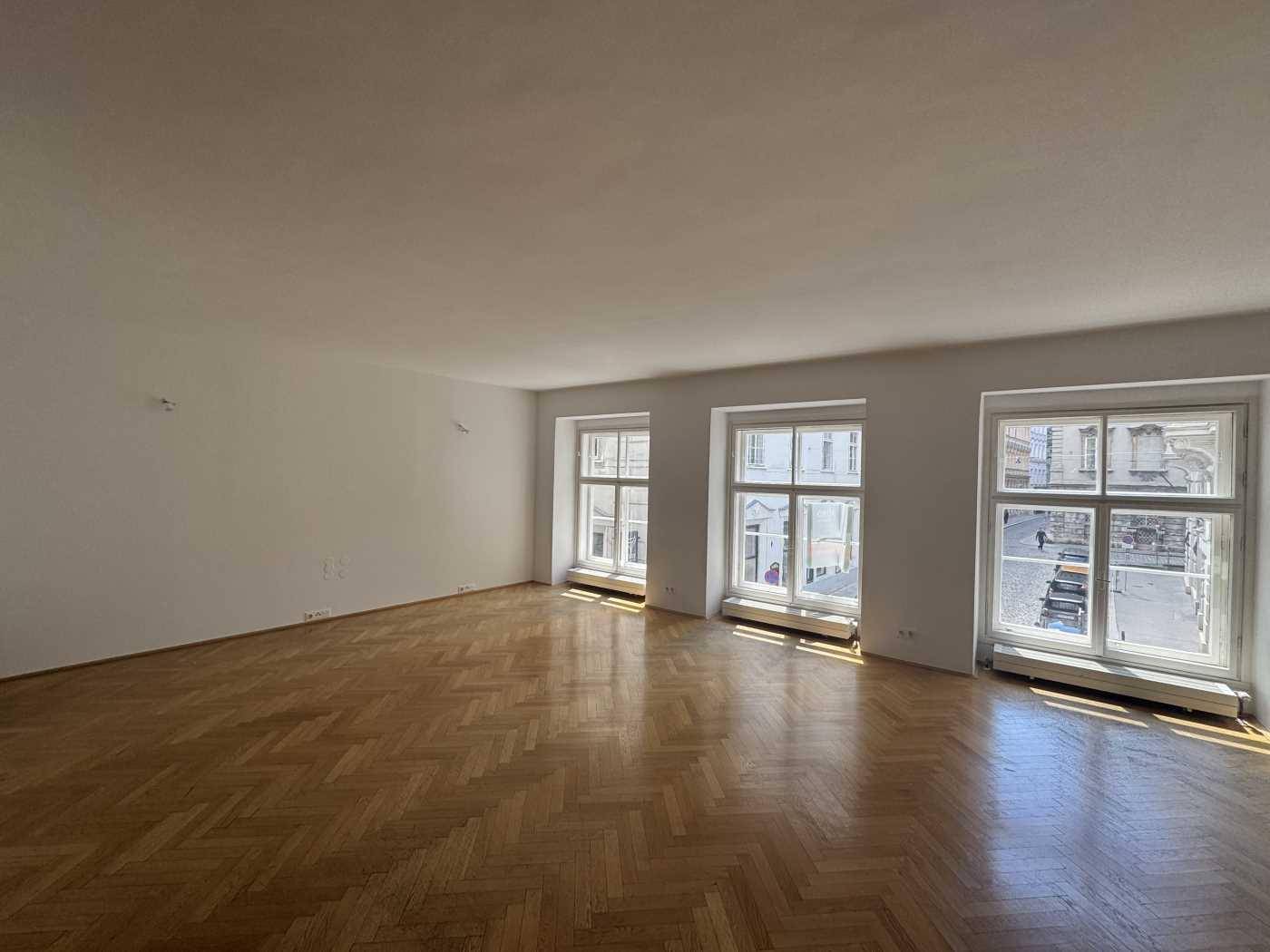 Großer, heller Raum mit Fischgrätparkett und drei hohen Fenstern, die den Blick auf die Stadt ermöglichen.