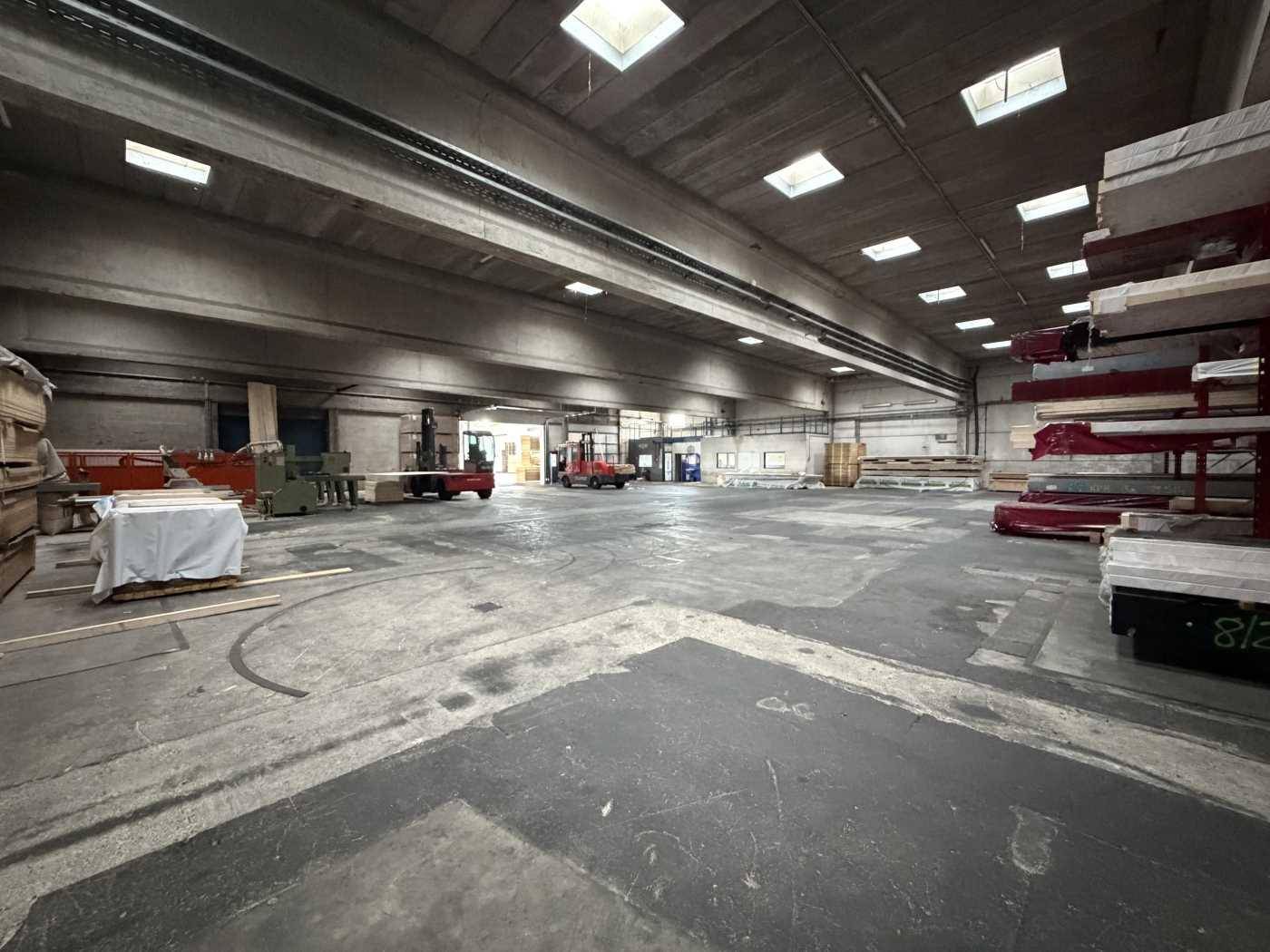 Weitläufige Lagerhalle mit viel freiem Raum, Betonboden und Gabelstaplern im hinteren Bereich.