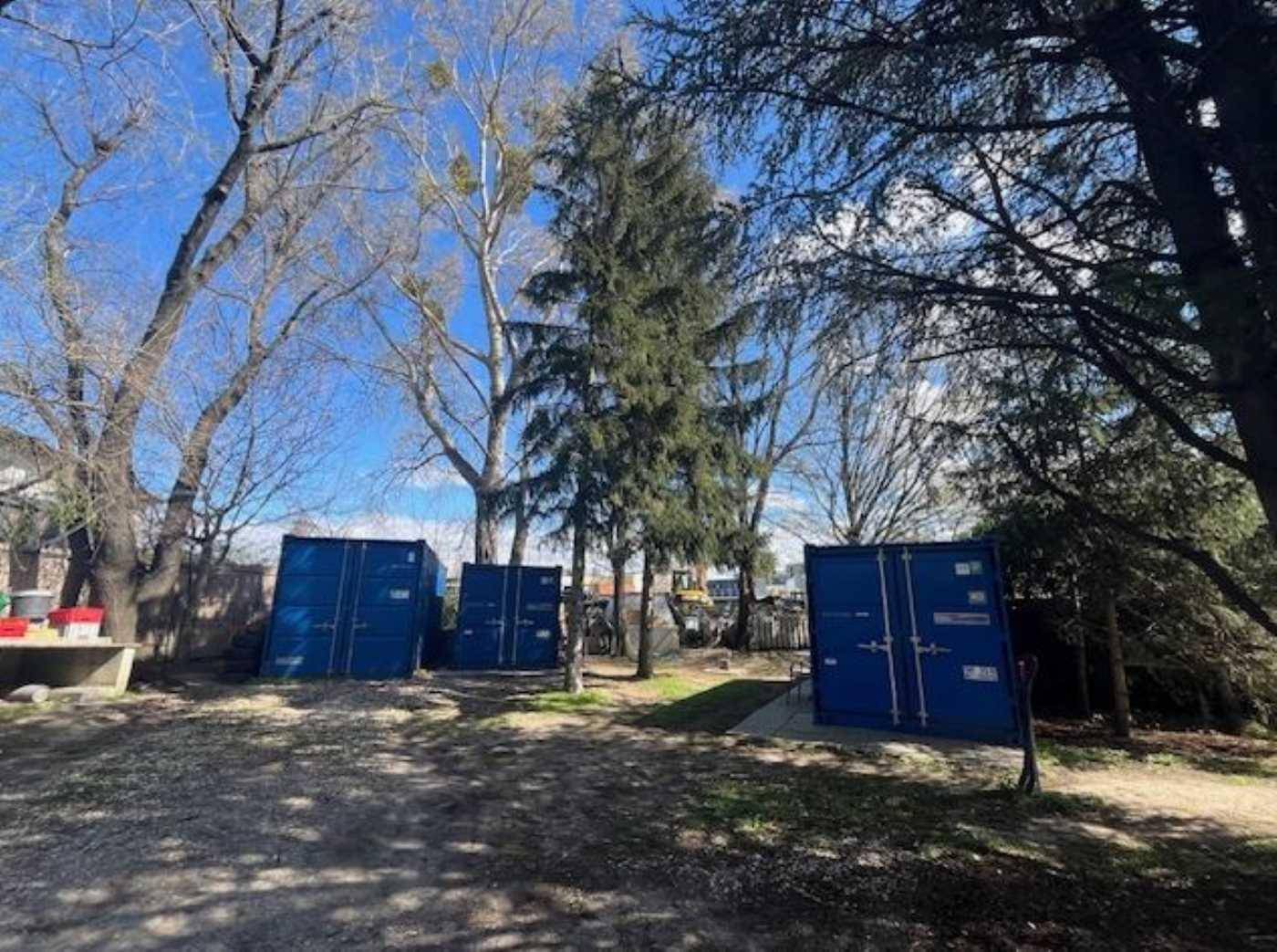 Außenbereich mit Bäumen, Gras und drei blauen Lagercontainern unter klarem Himmel.