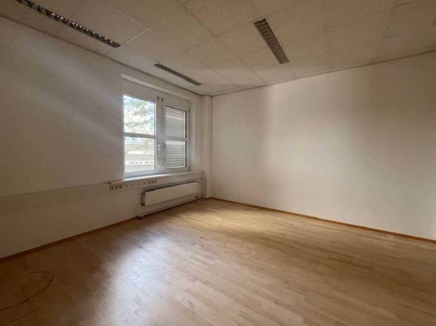 Heller Büroraum mit Holzboden, weißen Wänden und einem Fenster mit Jalousien, ideal für Einzelbüro.