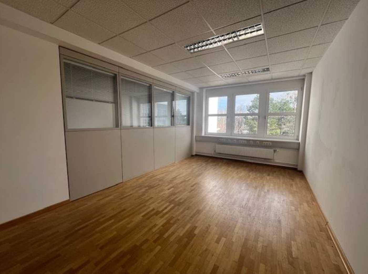 Büroraum mit Holzboden, großen Fenstern und einer Trennwand mit Glaspaneelen für mehr Privatsphäre.
