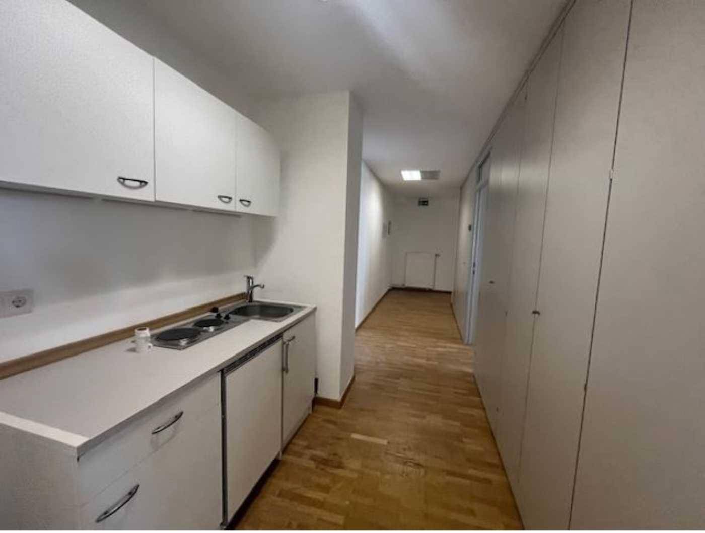 Kleine Küchenzeile in einem Flur mit weißen Schränken, Spüle und zwei Kochplatten, praktisch und kompakt.