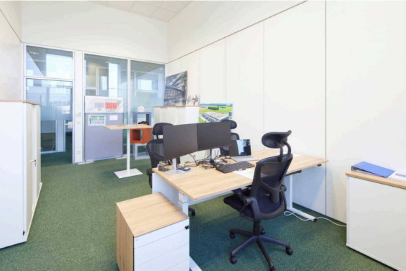 Modernes Büro mit grünem Teppichboden, ergonomischem Schreibtisch und Glaswänden zu weiteren Büroräumen.