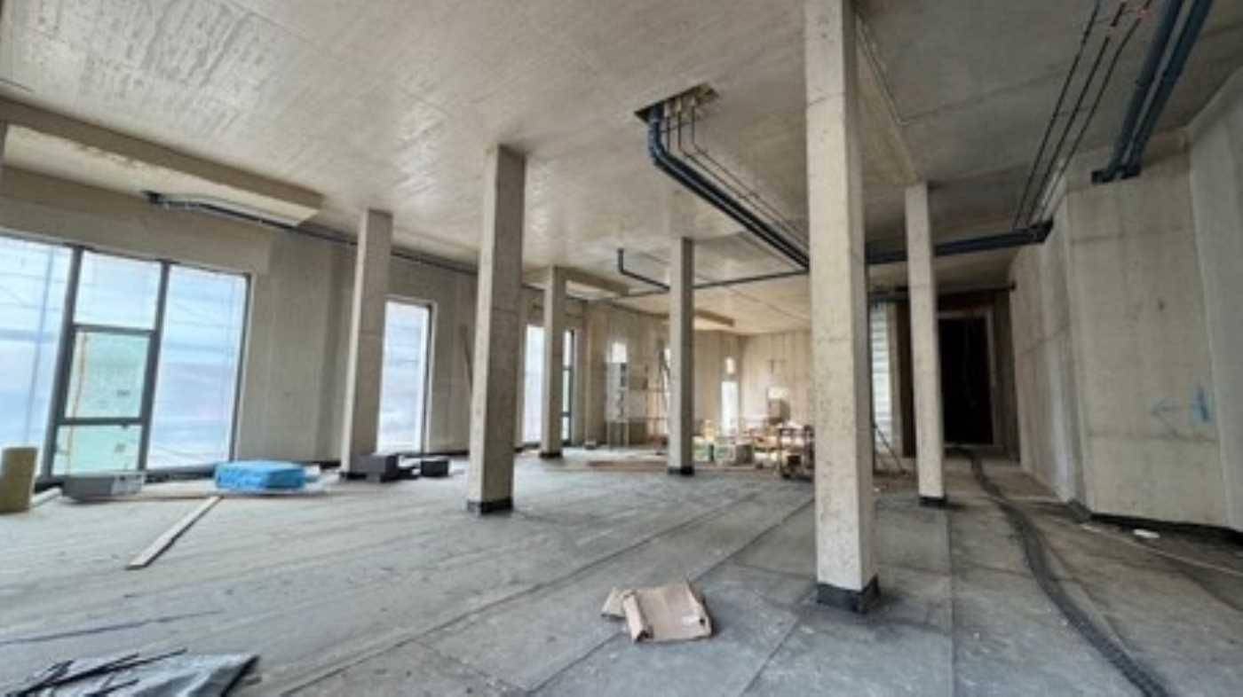 Innenansicht eines Rohbaus mit Betonstützen, großen Fenstern und sichtbaren Installationen während der Bauphase.
