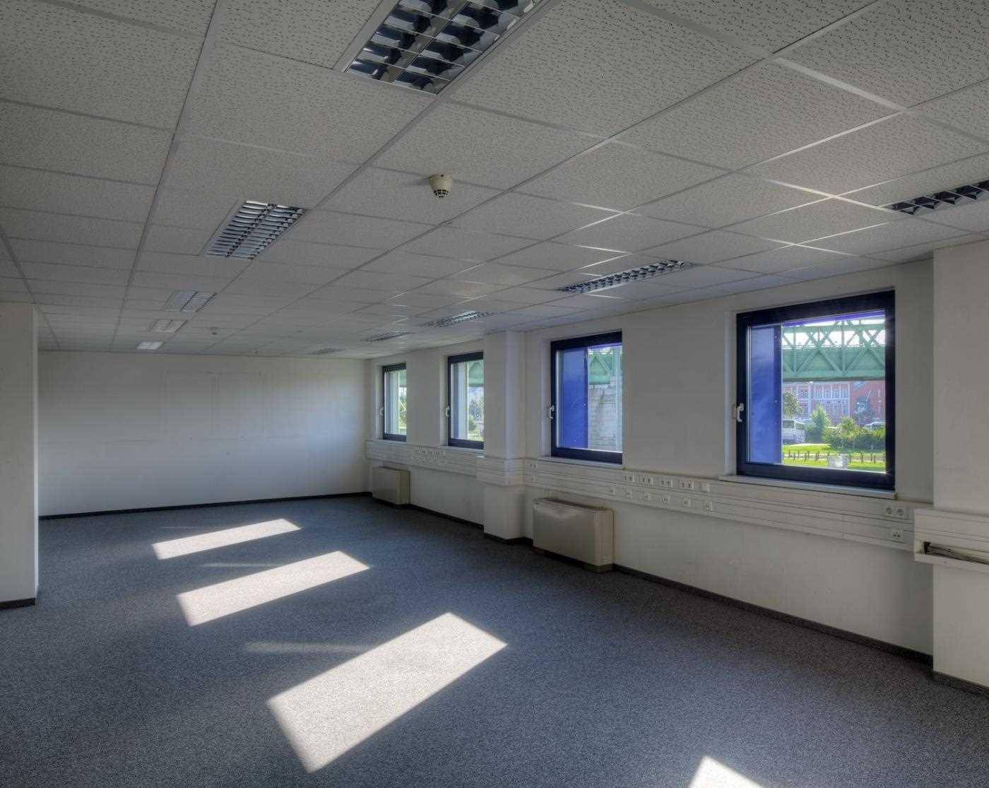 Leeres Büro mit grauem Teppichboden, weißen Wänden und Rasterdecke, durch die Fenster fällt Tageslicht.
