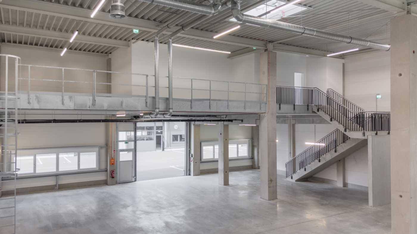 Geräumige, helle Halle mit hohen Decken, Betonboden, einer Galerie und einer Treppe, ideal für Lager oder Produktion.