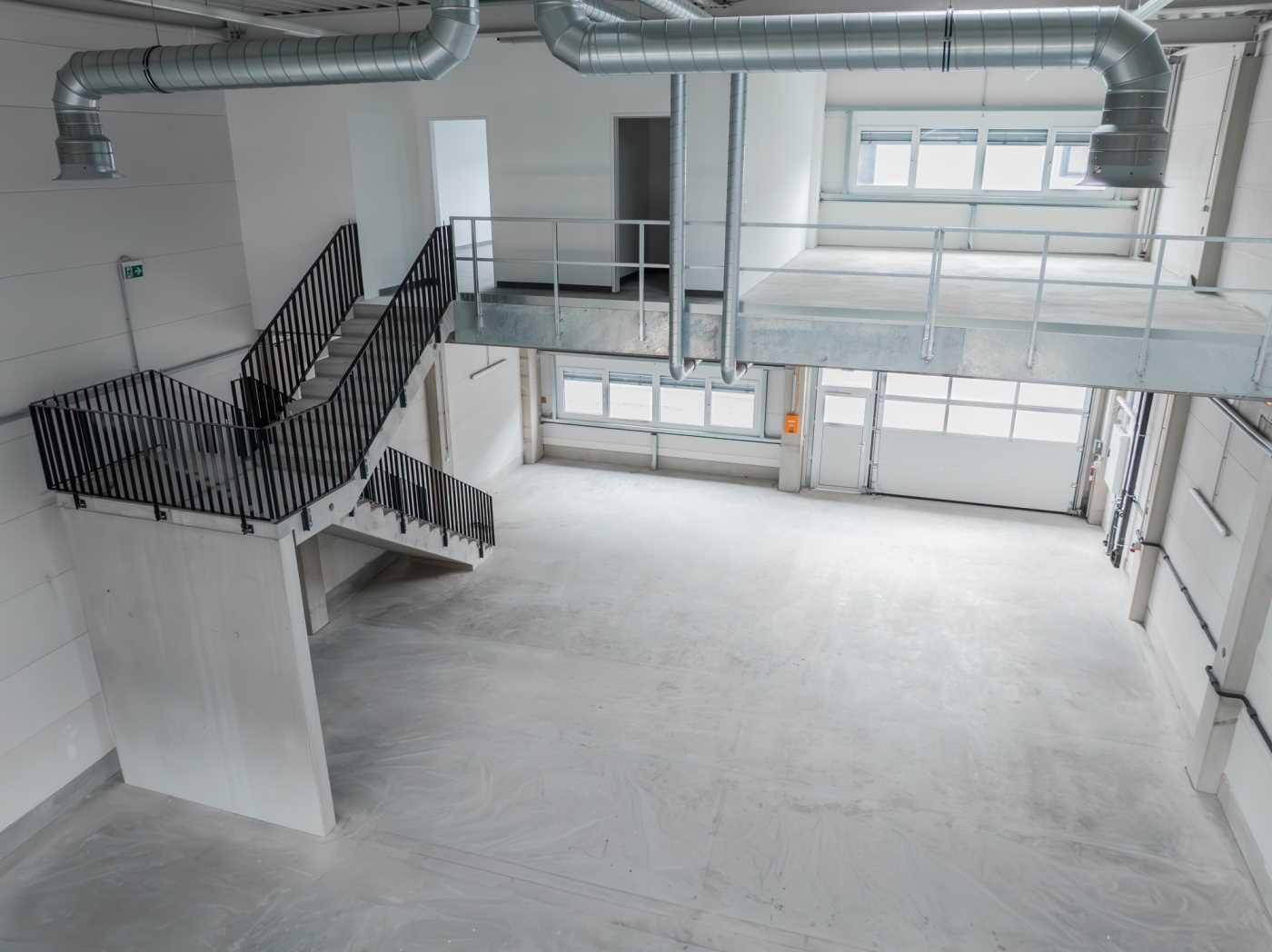 Geräumige, helle Halle mit hohen Decken, Betonboden, einer Galerie und einer Treppe, ideal für Lager oder Produktion.