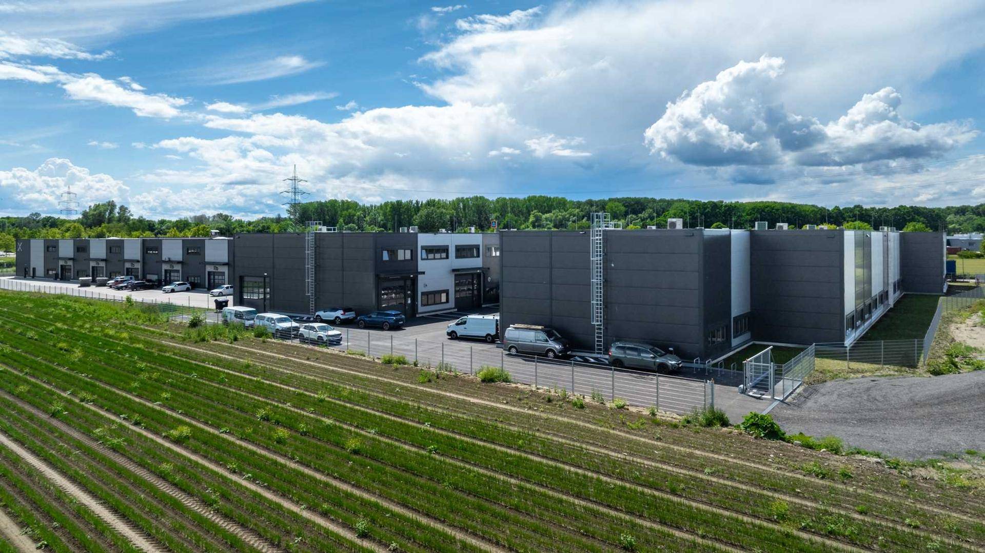 Weitläufige Ansicht eines modernen Gewerbeparks mit mehreren Gebäuden und angrenzenden Feldern unter blauem Himmel.