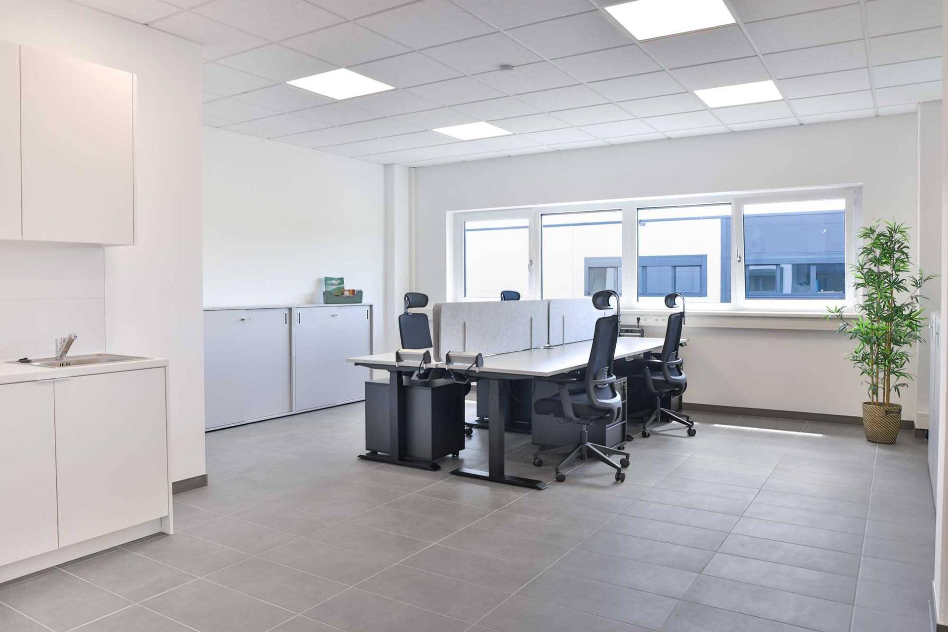 Helles, modernes Büro mit mehreren Arbeitsplätzen, großen Fenstern und einer kleinen Küchenzeile.