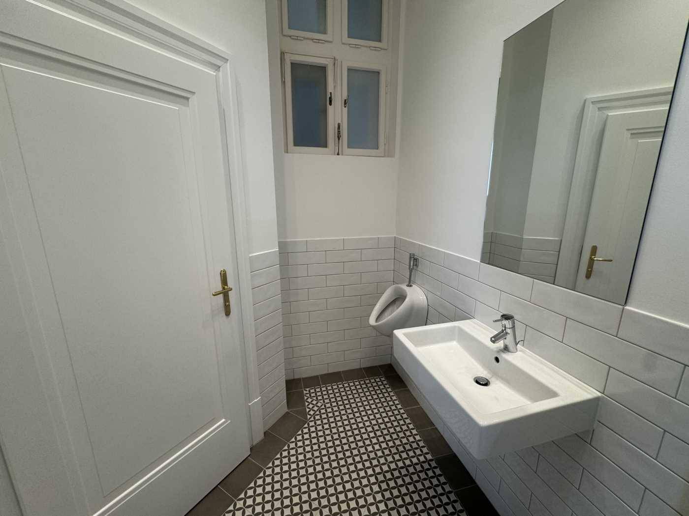 Modernes Gäste-WC mit Urinal, Waschbecken, Spiegel und Fenster, hell gefliest und sauber.