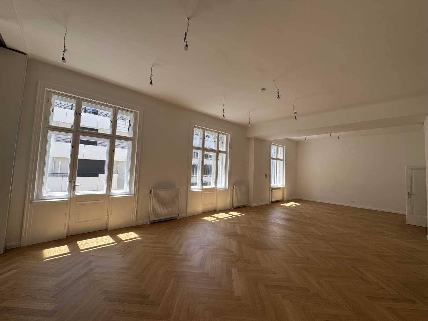 Großer, leerer Büroraum mit Fischgrätparkett und vielen Fenstern, die viel Tageslicht hereinlassen.