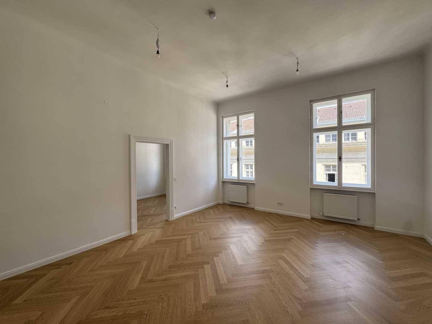 Geräumiger, heller Raum mit Fischgrätparkett und hohen Fenstern, bietet viel Tageslicht.