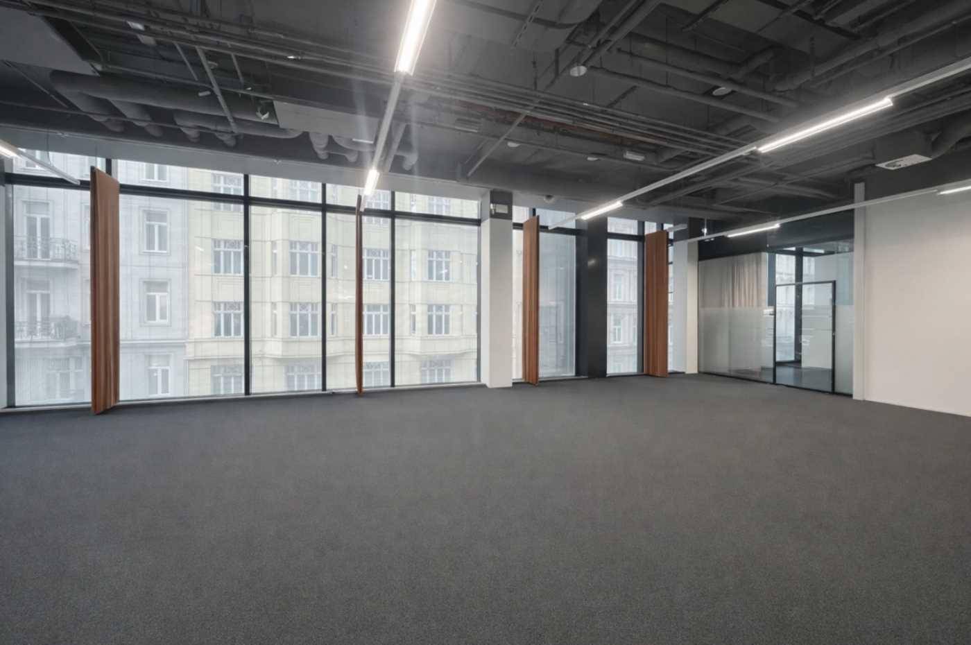 Heller, leerer Büroraum mit Teppichboden, großen Fenstern und Blick auf gegenüberliegende Gebäude.