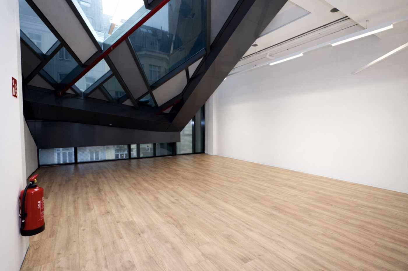 Leerer Büroraum mit Holzboden, großen Fenstern und einer markanten, schrägen Deckenkonstruktion.