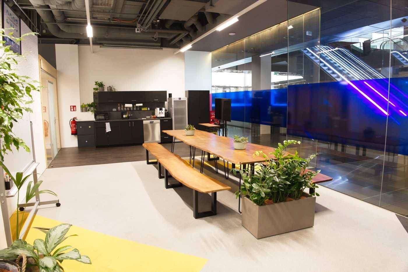 Moderne Büroküche mit Holztisch, Bänken und Pflanzen, angrenzend an einen Bereich mit Rolltreppen.
