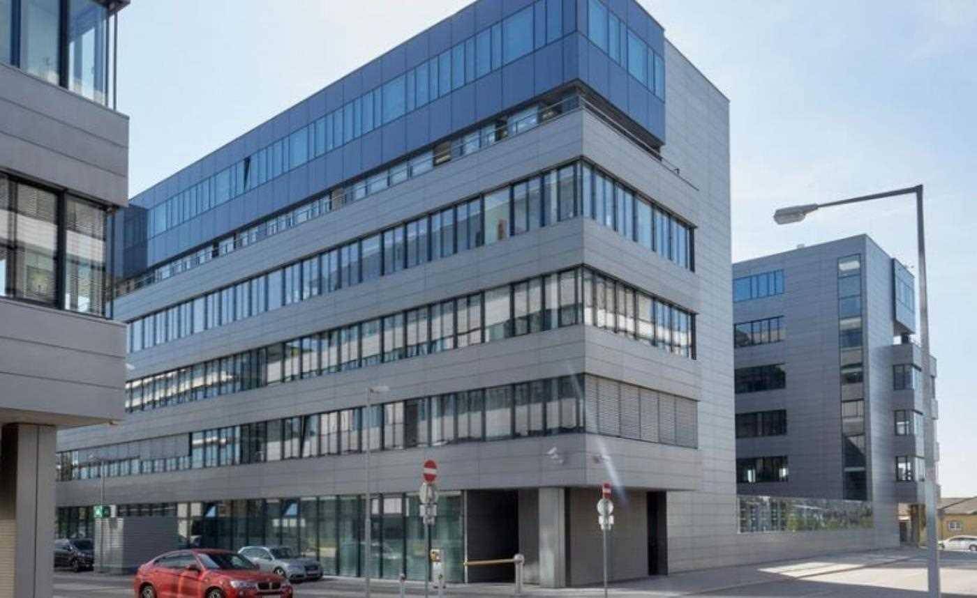 Modernes Bürogebäude an einer Straßenecke mit Glasfassade, Jalousien und angrenzenden Parkplätzen für Mitarbeiter und Besucher.