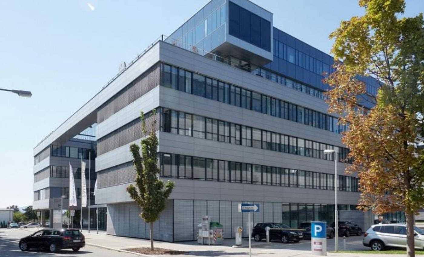 Modernes Bürogebäude mit großzügigen Fensterfronten, Jalousien und angrenzenden Parkmöglichkeiten unter blauem Himmel.