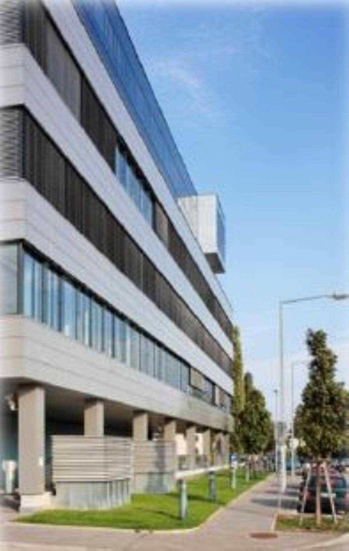 Seitenansicht eines modernen Bürogebäudes mit vielen Fenstern, Jalousien und gepflegten Grünflächen entlang des Gehwegs.