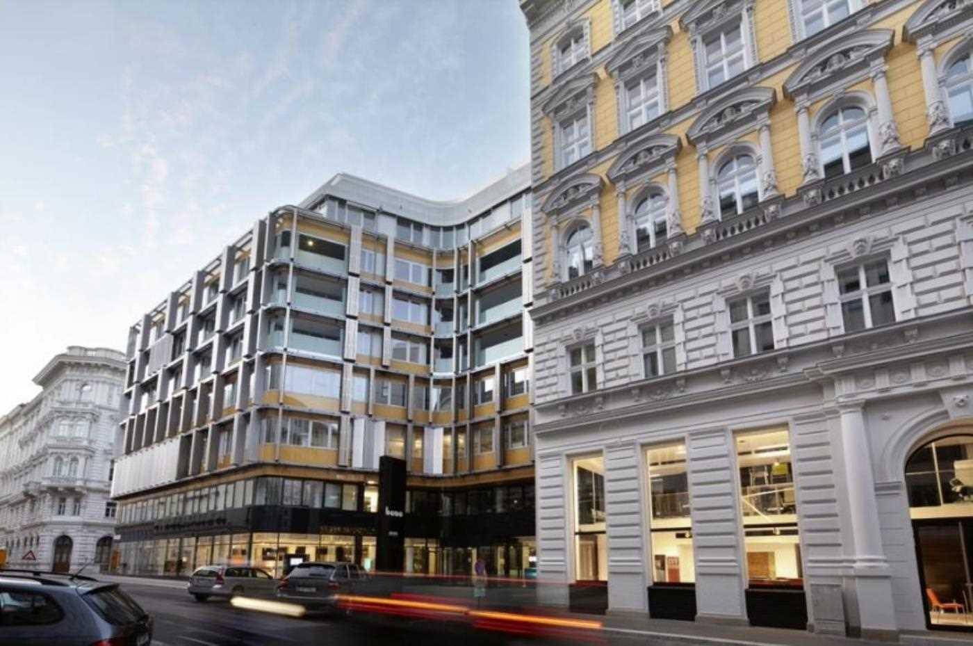 Straßenansicht des modernen Bürogebäudes neben einem traditionellen Altbau mit vorbeifahrenden Autos.