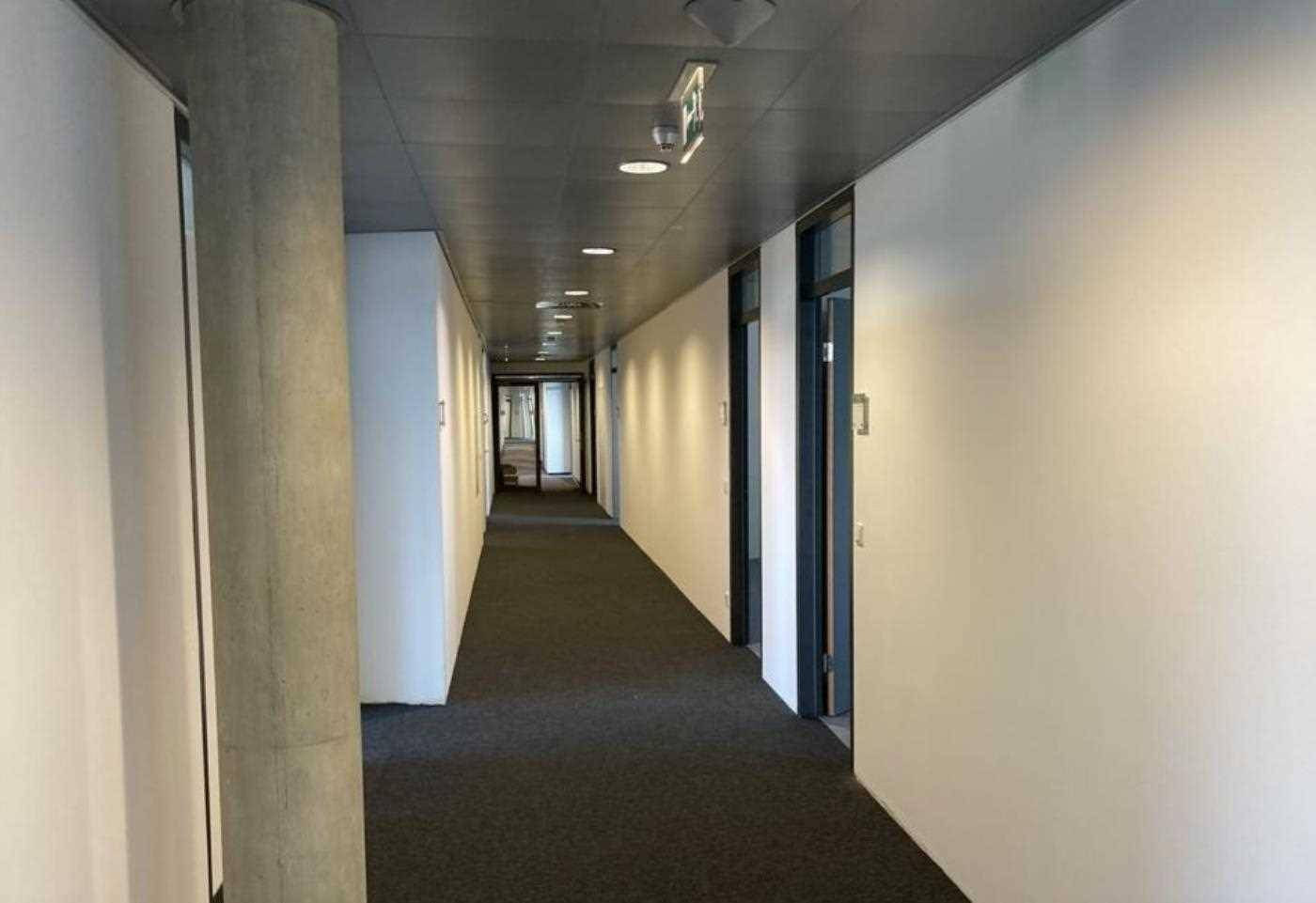 Langer Bürogang mit weißen Wänden, einem Betonpfeiler und dunklem Teppichboden.
