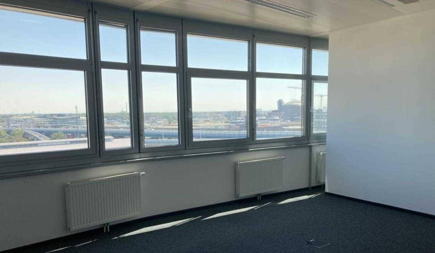 Leeres Büro mit großen Fenstern, Heizkörpern und Teppichboden, bietet einen weiten Stadtblick.